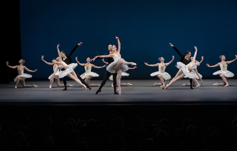 Some dancers in white tutus knee while others are partnered by male dancers in black.